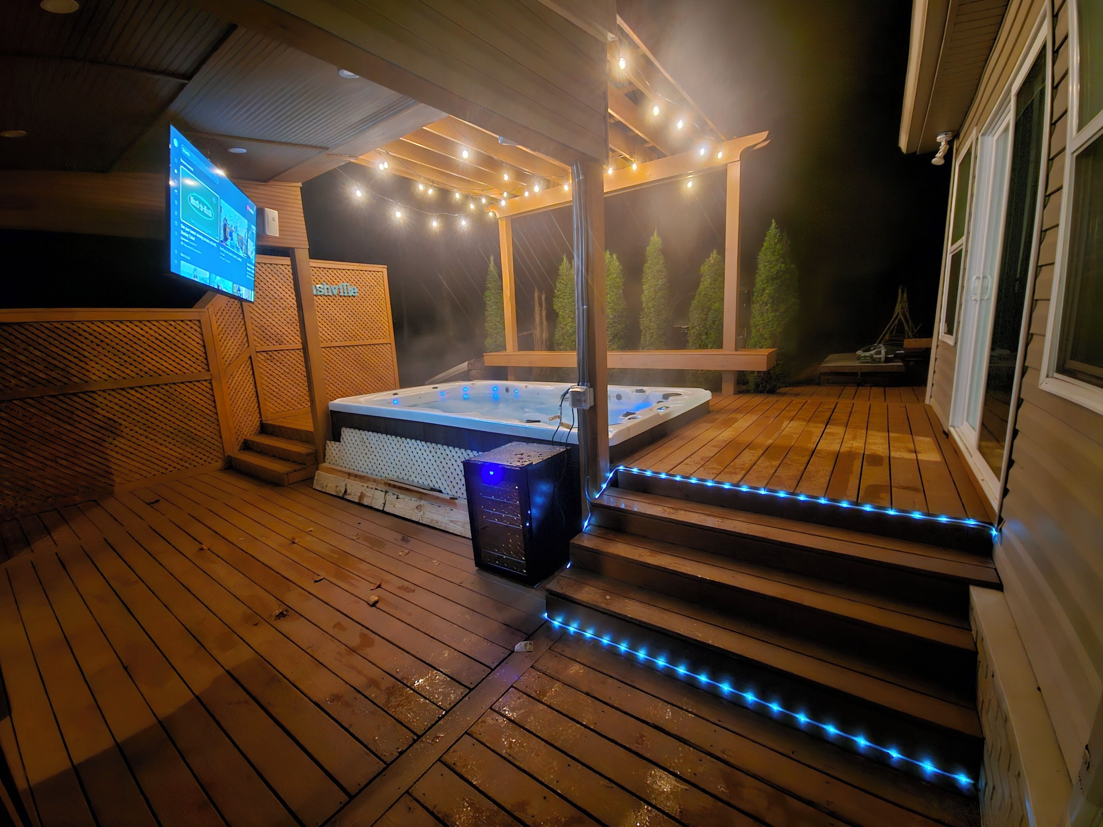 Eine Holzterrasse mit Whirlpool, Lichterketten und einem digitalen Bildschirm.