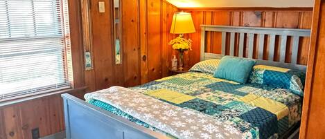 A bedroom with a wooden headboard, a bed with a patterned quilt, a bedside lamp, and a window with blinds.