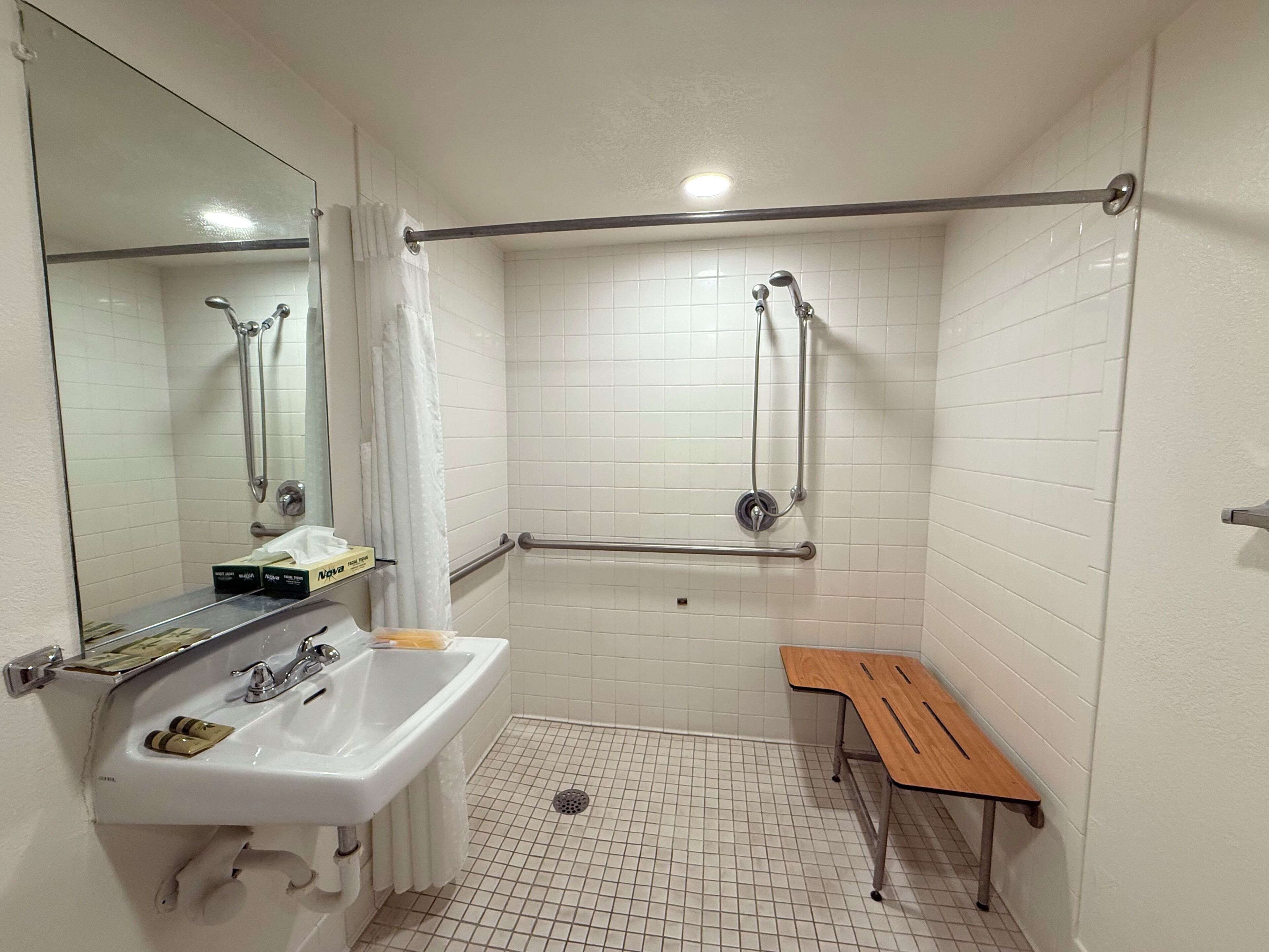 A bathroom with a sink, mirror, shower, grab bars, and a bench.