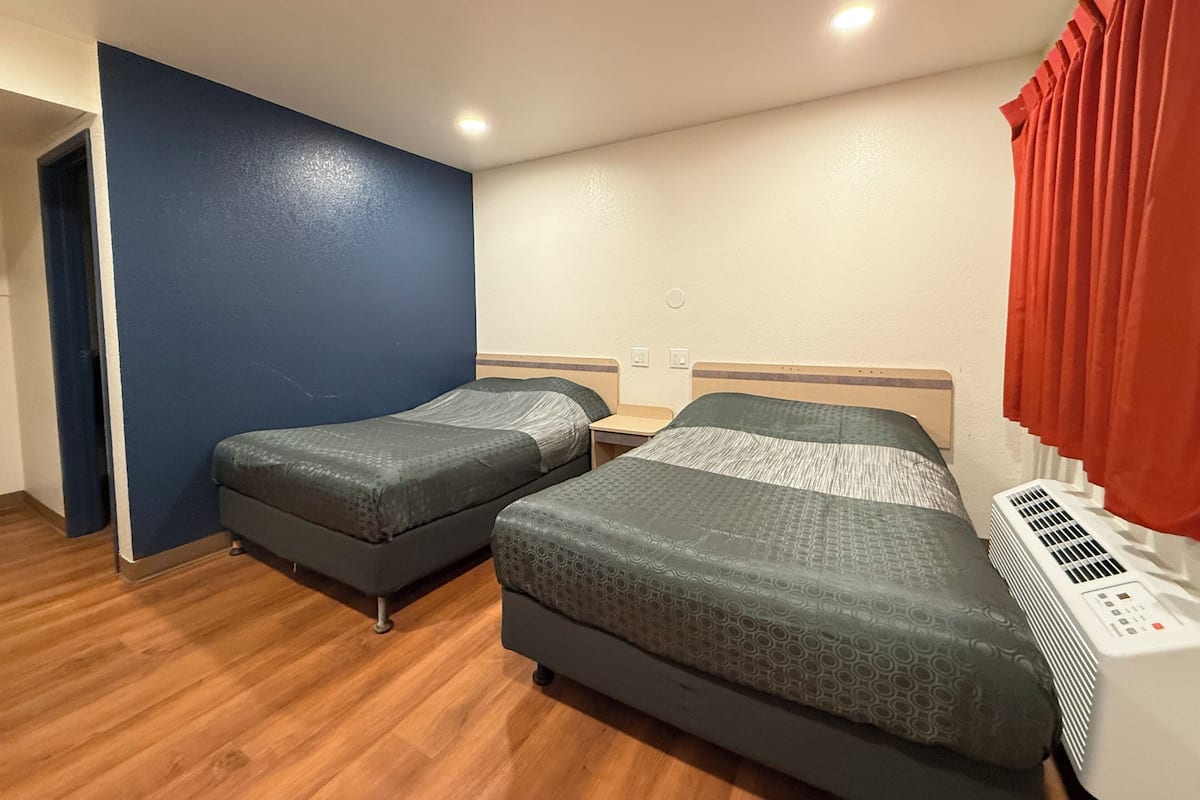 A hotel room with two beds, a wooden floor, a blue wall, a white ceiling, and a red curtain.