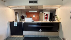 Una zona de recepció amb taulell, ordinador, extintor, rellotge i paperera.