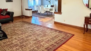 A spacious living room with a large patterned rug, a dining area with a white table and chairs, and a hallway leading to another room.