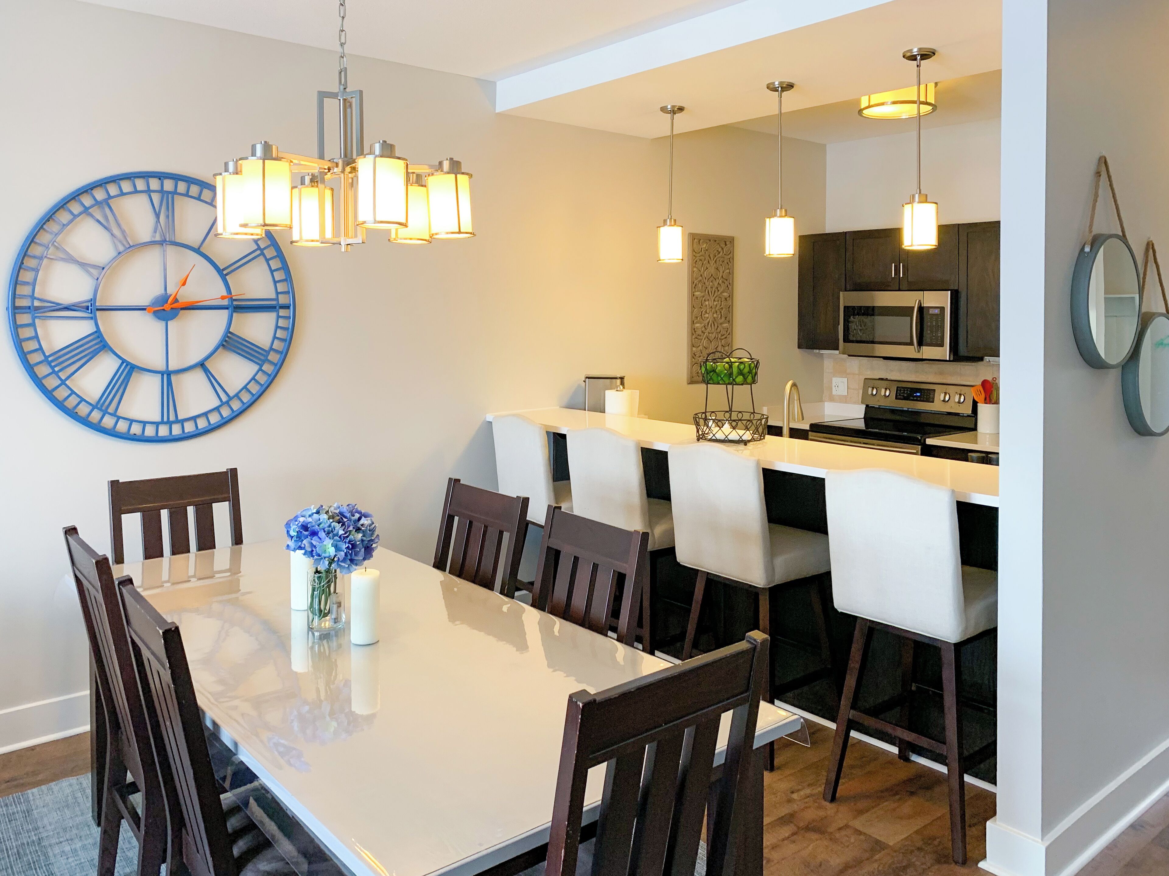 A modern dining area with a large wall clock, a chandelier, and a kitchen in the background.