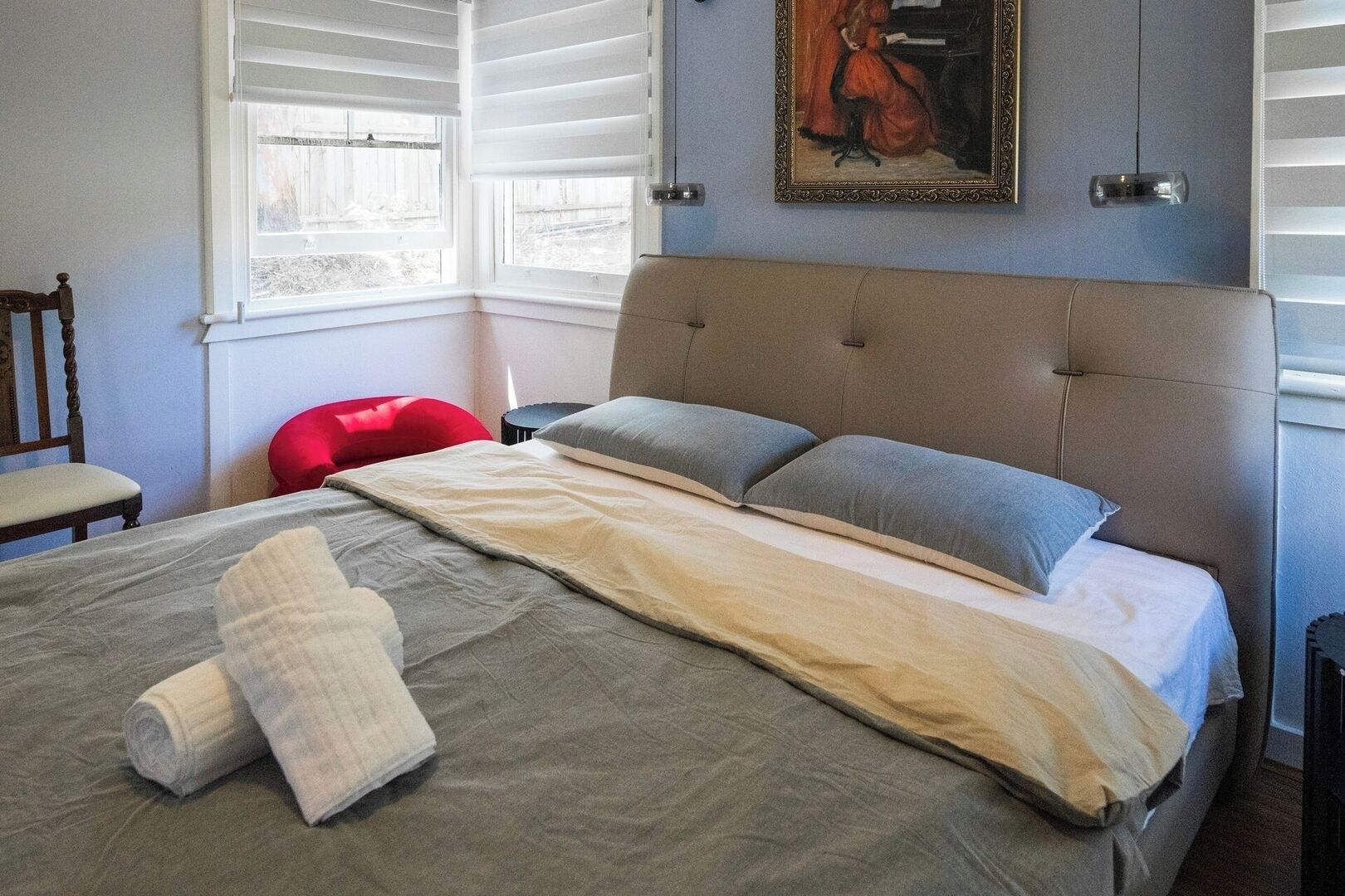 Un dormitorio con cama, almohadas, manta, una silla y un cuadro en la pared.