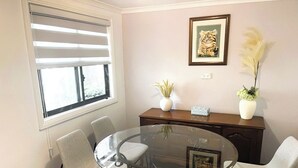 Un salón de conferencias con una mesa de vidrio rodeada de sillas, una ventana con persianas, un cuadro en la pared y dos plantas en macetas sobre un mueble de madera.