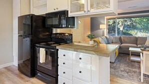 Eine moderne Küche mit schwarzem Kühlschrank, Herd und Mikrowelle. Es gibt einen Essbereich mit Tisch und Stühlen sowie ein Wohnzimmer mit Sofa und einem großen Fenster.