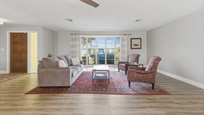 Uma sala de estar com piso de madeira, ventilador de teto, sofá, mesa de centro e duas poltronas estampadas.