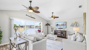 A spacious living area with a large window, ceiling fans, a television, and a view of the water. - Gulf Access, Heated Pool, Bikes- Coastal Breeze - Roelens Vacations (Cape Coral)