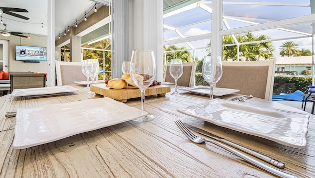 A dining table set with white plates, cutlery, and wine glasses, with a wooden board holding bread and a bottle. - Gulf Access, Heated Pool, Bikes- Coastal Breeze - Roelens Vacations (Cape Coral)