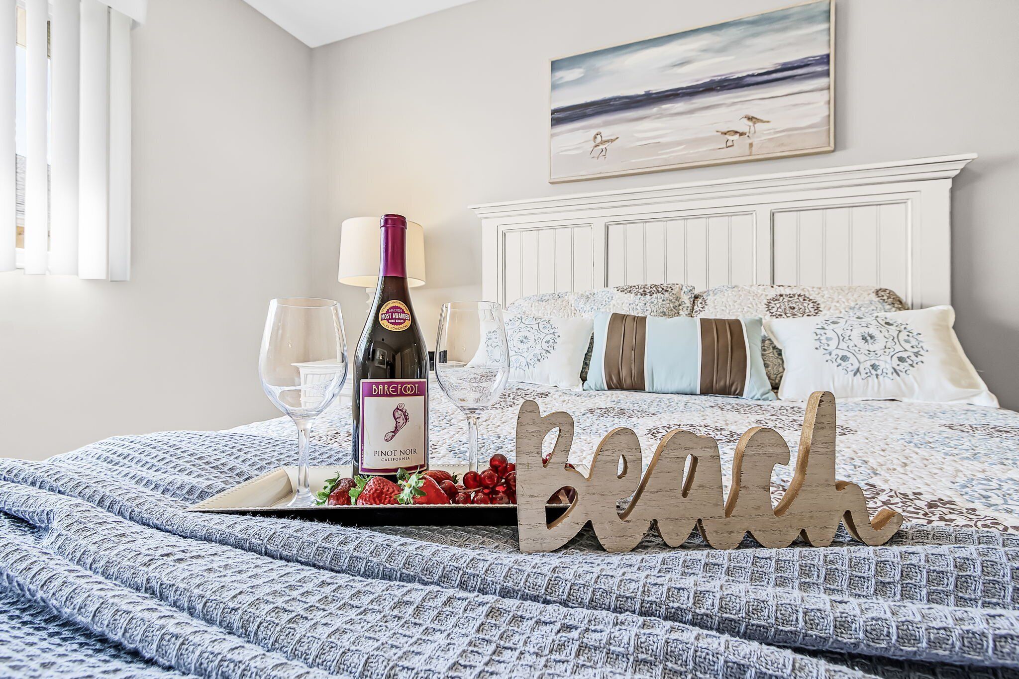 Una cama impecable con un cartel que dice "Beach", una botella de vino y copas sobre una bandeja con fresas.