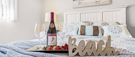 Una cama impecable con un cartel que dice "Beach", una botella de vino y copas sobre una bandeja con fresas.