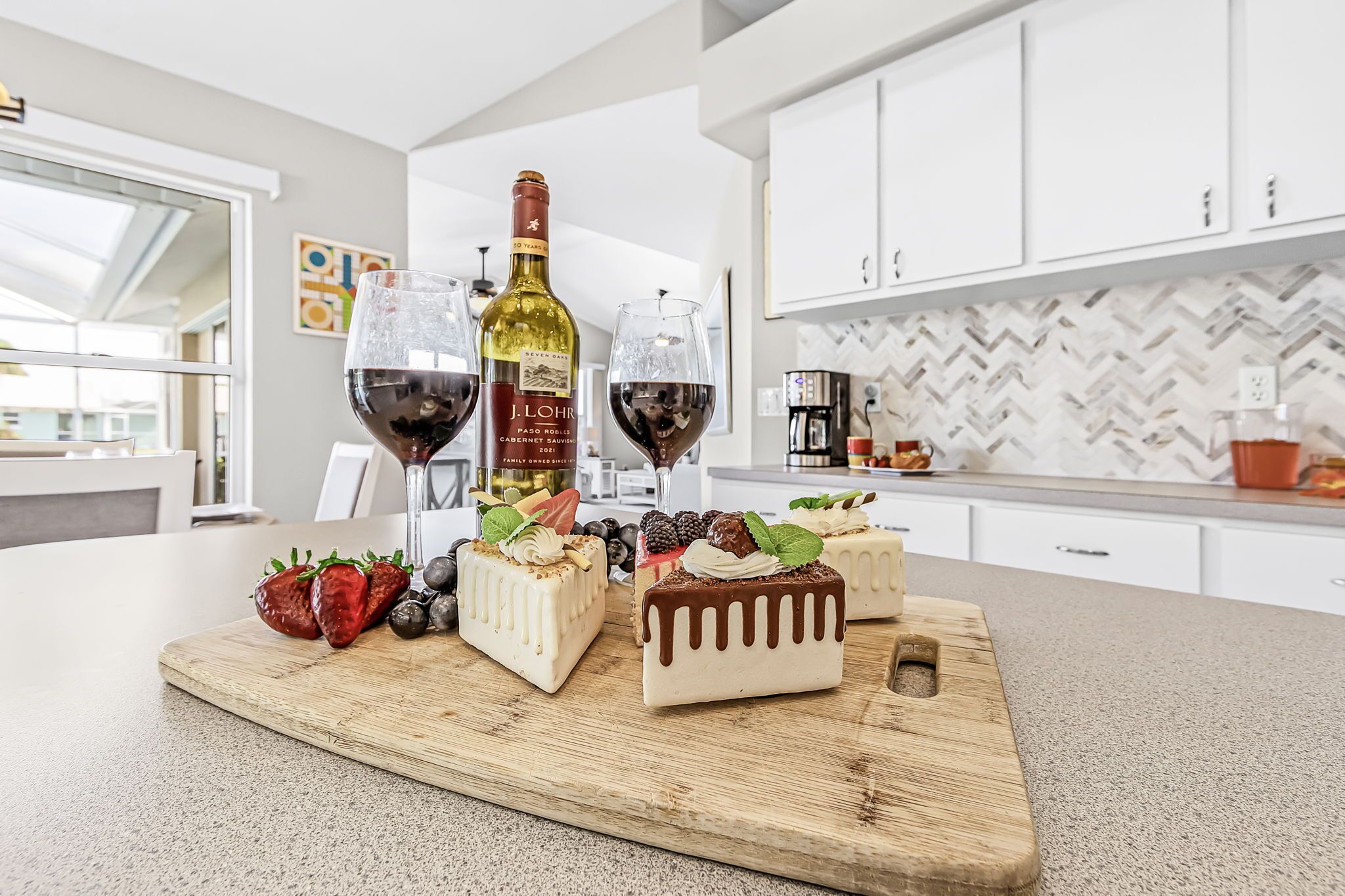 A kitchen counter with three square cakes, two glasses of red wine, and a bottle of wine.