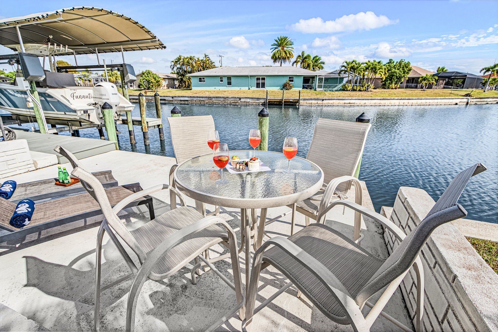 A table with chairs and a drink, overlooking a body of water with a house and palm trees in the background.