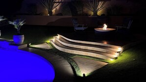 Un patio al aire libre iluminado por la noche con un brasero, dos sillas y una piscina con forma curva.