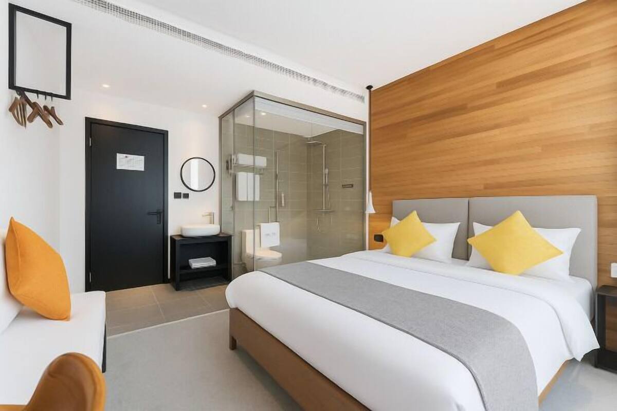 Una habitación de hotel moderna con una cama grande, ducha con mampara de vidrio, un panel de madera decorativo y una mesita de noche pequeña.
