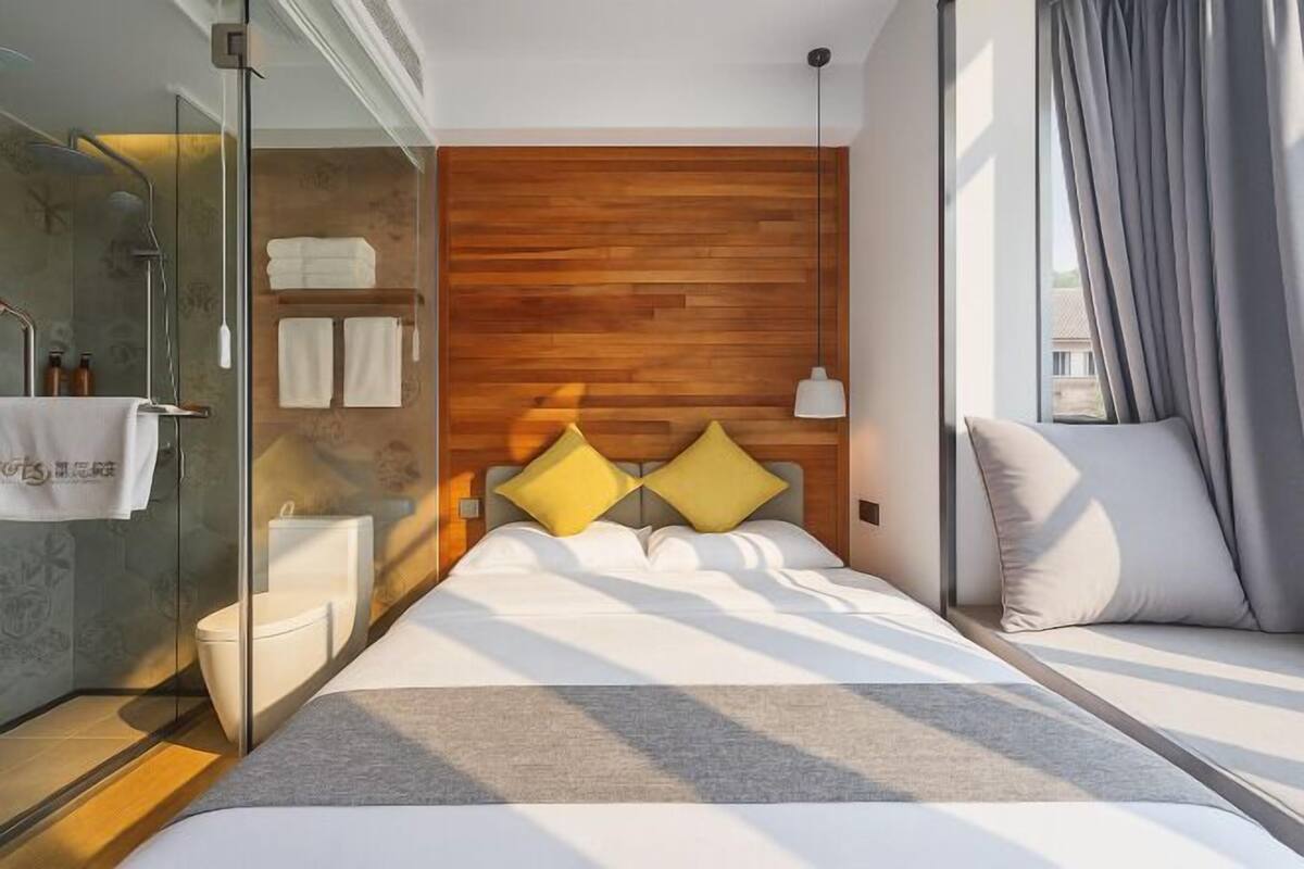 Una habitación de hotel moderna con cabecera de madera, cerramiento de ducha de vidrio, cama con ropa de cama blanca y vistas al exterior.