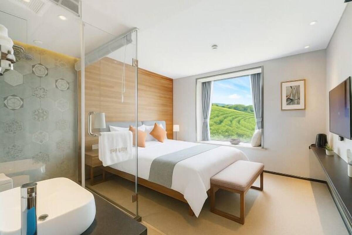Una habitación de hotel moderna con una cama grande, cabina de ducha de vidrio, vista a campos verdes y un televisor de pantalla plana.