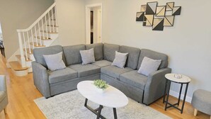 Uma sala moderna com um sofá modular cinzento, uma mesa de centro redonda, uma pequena mesa lateral e uma escada com guarda-corrimão branco.