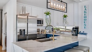 A modern kitchen with a stainless steel refrigerator, a built-in microwave, and a coffee machine. There are hanging plants and a chandelier above the island. - Cayuga Lake Holiday House - Waterfront w/ Sunset Views (Ledyard)