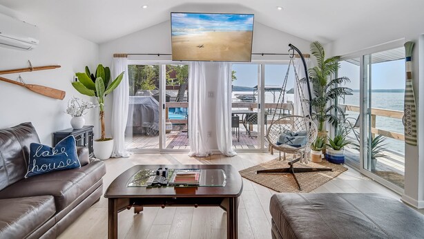 A spacious living room with a large TV, leather sofas, a coffee table, and a view of the beach through large windows. - Cayuga Lake Holiday House - Waterfront w/ Sunset Views (Ledyard)