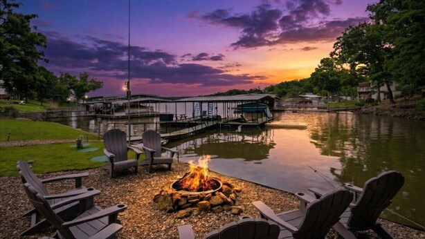 A lakeside setting with a fire pit and Adirondack chairs, a dock, and a building with a covered walkway.
