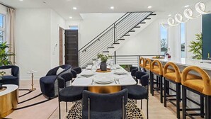A modern dining area with a central table set for six, flanked by black and mustard chairs, a staircase, and a view of the outdoors.