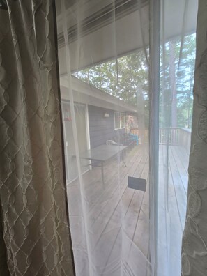 A view through a window with a patterned curtain, a patio area with a table and chairs, and a wooden deck.