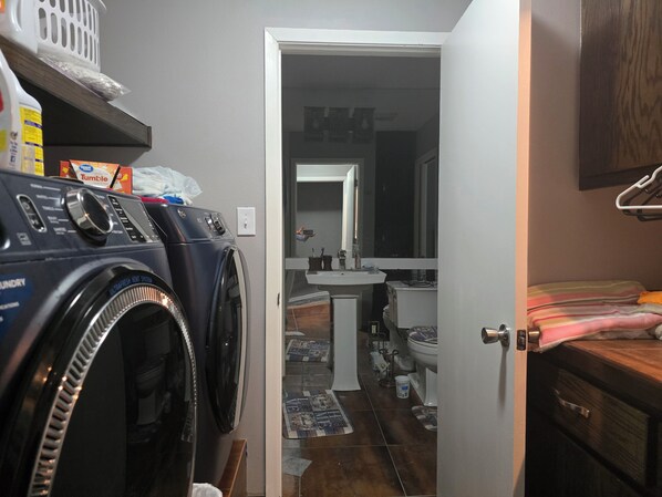 A laundry area with a front-loading washer and dryer, a bathroom with a sink and toilet, and a bed with a striped blanket.
