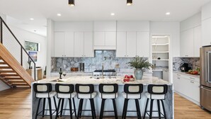 A modern kitchen with a marble island, bar stools, a stainless steel refrigerator, and a wooden staircase. - Sundown Social by Avantstay Lake Austin Views Pool, Firepit, Deck + Game Room A+ Designer Style (Austin)
