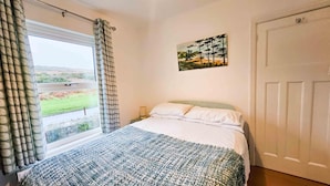 A bedroom with a bed, a window with curtains, a painting on the wall, and a door. - BrynView - 3 Bedroom Holiday Home - Reynoldston (Swansea)