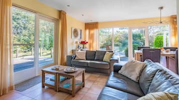 A spacious living area with a sofa, a coffee table, and a dining area with a table and chairs. Large windows provide a view of the outdoors.