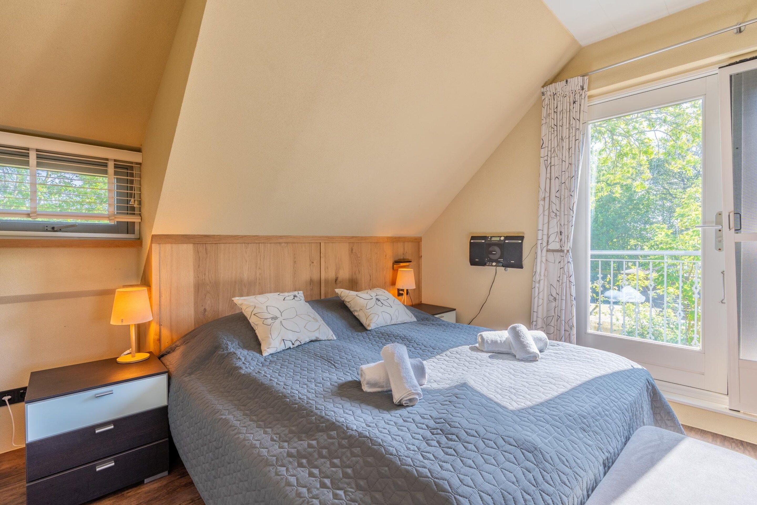 A bedroom with a large bed, a nightstand, a lamp, and a view of the outdoors through a window.