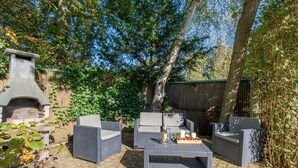 An outdoor patio with wicker furniture, a stone fireplace, and a wooden table set with candles and snacks. - Very Tastefully Furnished Country House for 4 People (Den Burg)