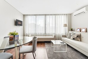 Ein modernes Wohnzimmer mit einem Glastisch, einer Couch und einem Couchtisch. Auf dem Tisch steht eine Pflanze, auf der Couch eine Lampe und an der Wand ist ein Fernseher angebracht.