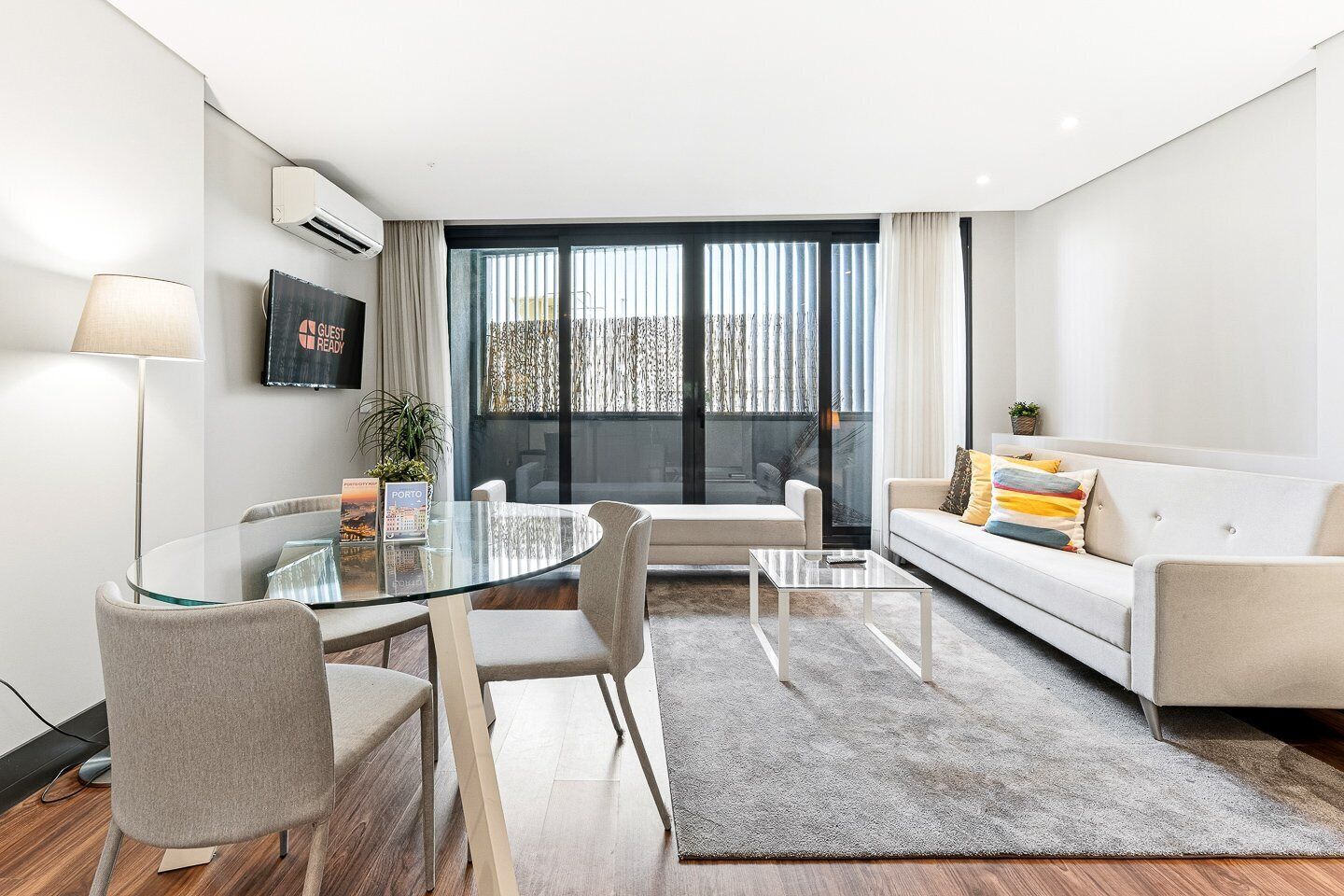 A modern living room with a glass dining table, a sofa, and a coffee table. There is a TV mounted on the wall and a window with curtains.