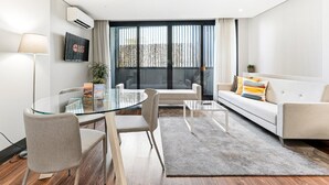 A modern living room with a glass dining table, a sofa, and a coffee table. There is a TV mounted on the wall and a window with curtains. - GuestReady - Ideal Porto stay near Trindade GF-B (Porto)