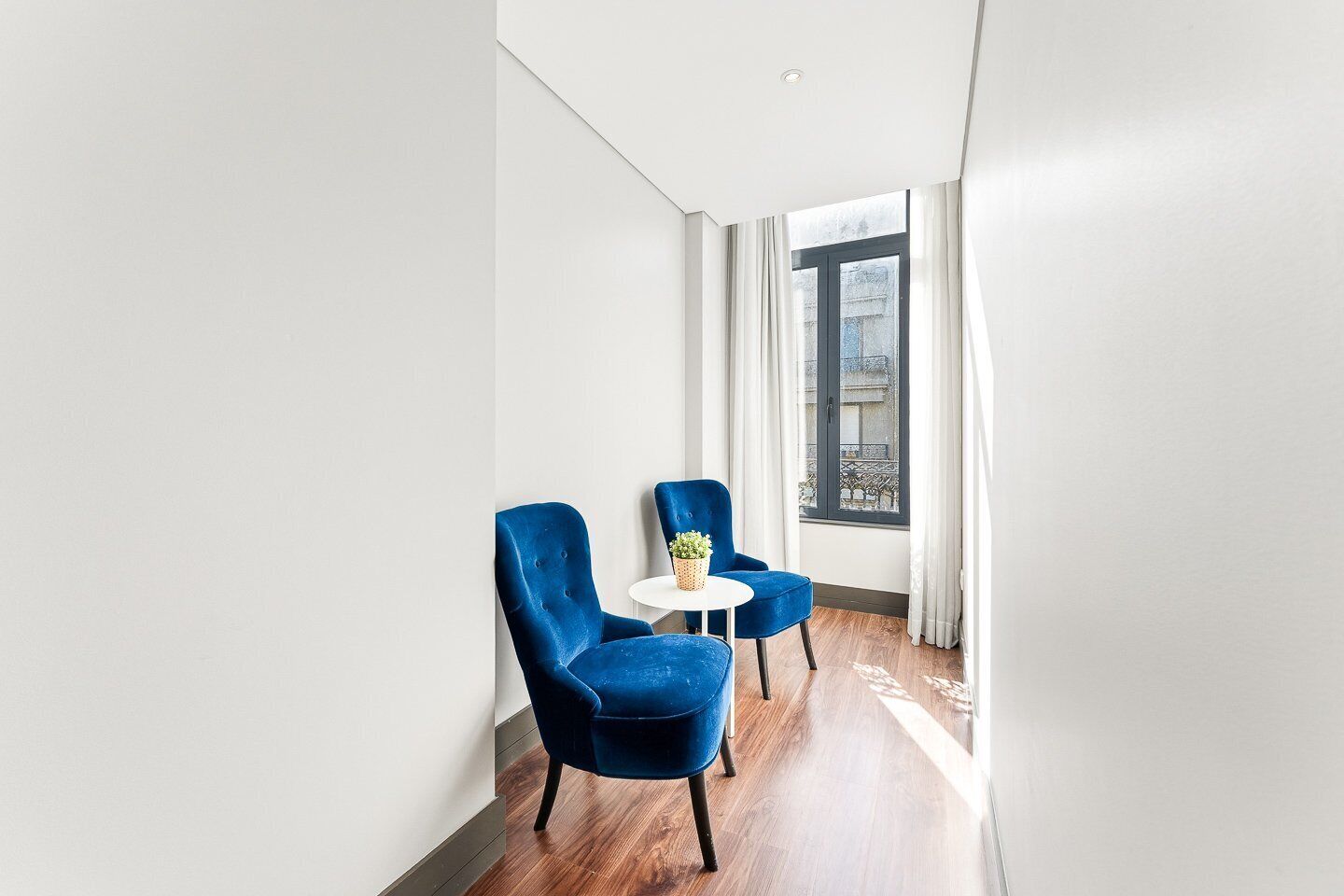 A narrow hallway with two blue armchairs and a small round table with a plant, leading to a window with a view of buildings.