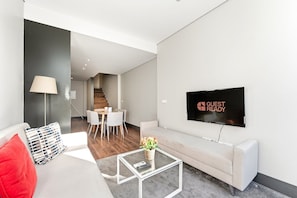 A modern living room with a sofa, a coffee table, a TV, and a dining area with a staircase in the background. - GuestReady - Ideal Porto stay near Trindade GF-F (Porto)