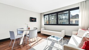 A modern living room with a round glass table, two grey chairs, a white sofa, and a coffee table. A flat-screen TV is mounted on the wall, and a window offers a view of buildings. - GuestReady - Ideal Porto stay near Trindade 2nd (Porto)