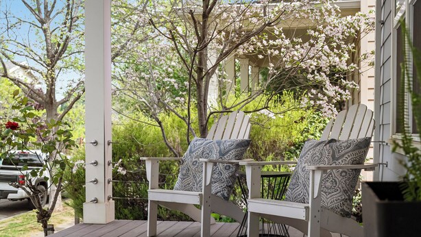 Um deck de madeira com duas cadeiras Adirondack, um corrimão branco e um alpendre com vista para um jardim e um edifício com colunas.