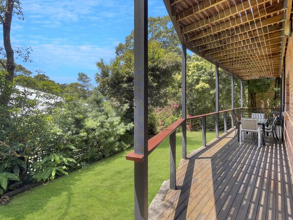 A wooden deck with a pergola, surrounded by lush greenery and a clear blue sky. - Relaxing 3-bedroom Retreat With Outdoor Seating (Lilli Pilli)