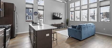 A modern kitchen with a central island, a dining area with a sofa, and large windows offering views of the outside.