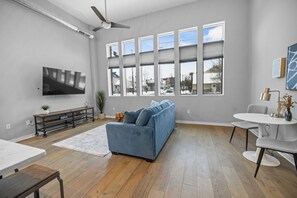 Ein modernes Wohnzimmer mit großem Fenster, Flachbildfernseher, einer Couch, einem Esstisch und einem Deckenventilator.