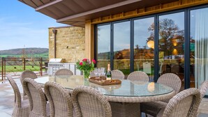 An outdoor dining area with wicker chairs and a glass table, a grill, and a flower arrangement. - Badgerwood - sleeps 12 guests  in 6 bedrooms (Winchcombe)
