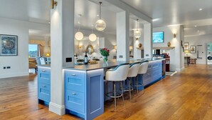 A modern kitchen with a central island, bar stools, pendant lighting, and a large window overlooking a garden. - Badgerwood - sleeps 12 guests  in 6 bedrooms (Winchcombe)