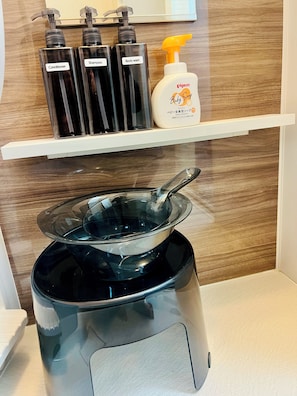 Kylpyhuoneessa on musta sähköinen vedenkeitin, valkoisella tasolla oleva lasikulho sekä hylly, jossa on kolme mustaa pulloa, joissa on etiketit: hoitoaine, shampoo ja suihkugeeli.