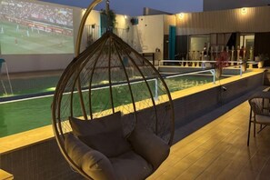 A hanging chair by a pool with a sports event on the screen, a bar area, and a view of the pool. - Al Rayyan Tower (Jeddah)