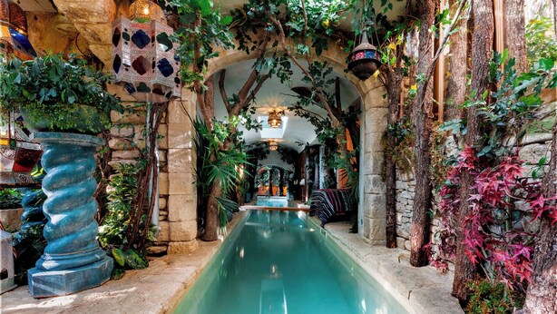 Une piscine intérieure aux murs et aux arcades en pierre, entourée d’une végétation luxuriante et de plantes suspendues.