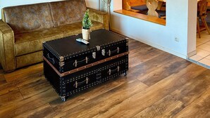 A living room with a brown leather sofa, a black suitcase coffee table with a plant, wooden flooring, a painting on the wall, and a view into the dining area through a window with curtains. - Vacation apartment no. 2 for 1 - 4 persons 59 sqm (Reit im Winkl)