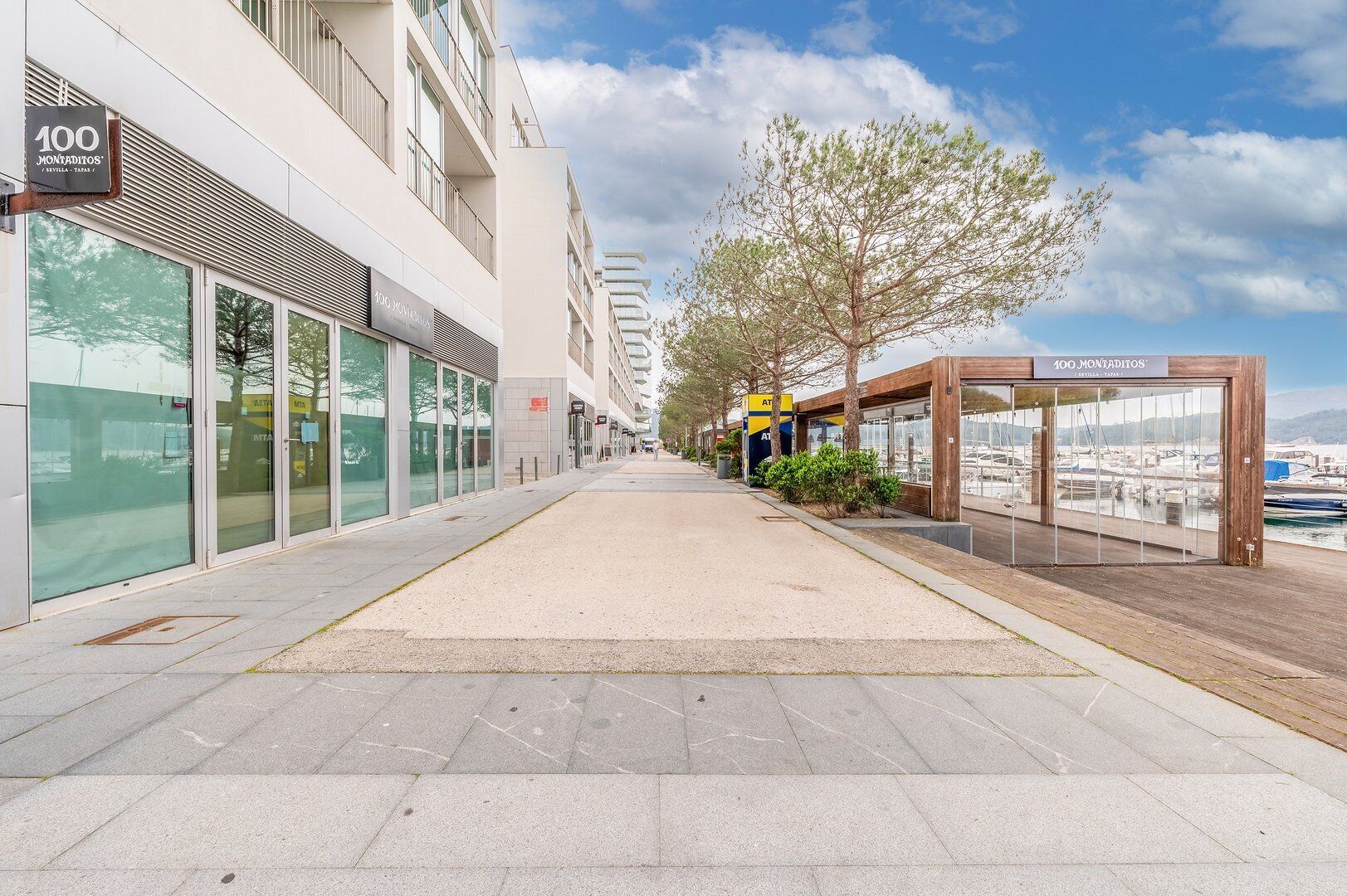 A modern building with large glass windows, a covered walkway, and a view of boats in the background.
